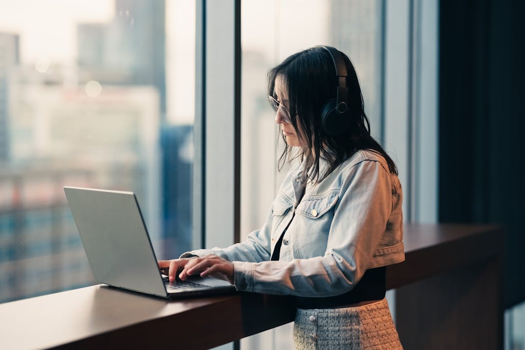 A woman wearing headphones works on her laptop in a high-rise building, looking out a large window with a city view researching s corp benefits and risks.