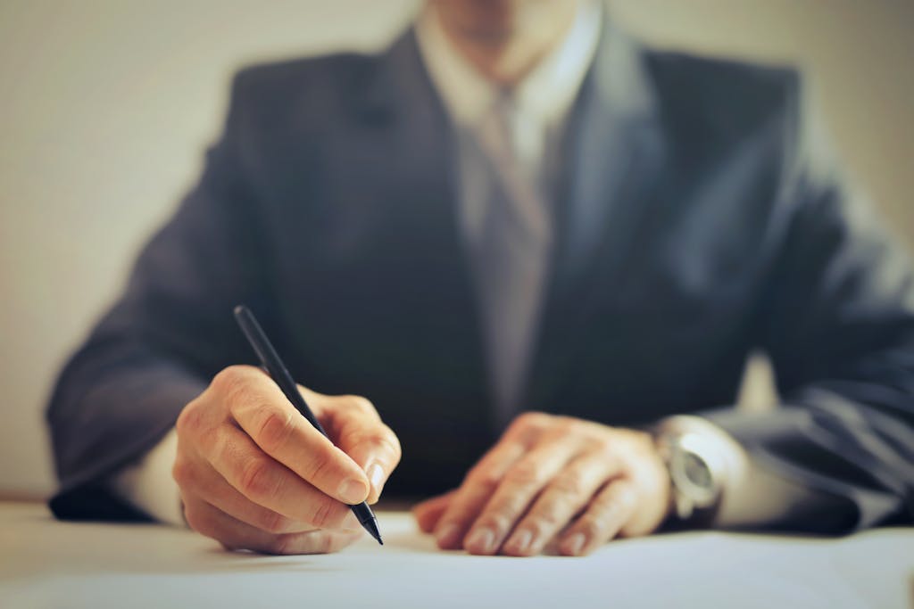 Faceless businessman in suit signing a contract at office desk, focused on reviewing the liquidated damages clauses. 