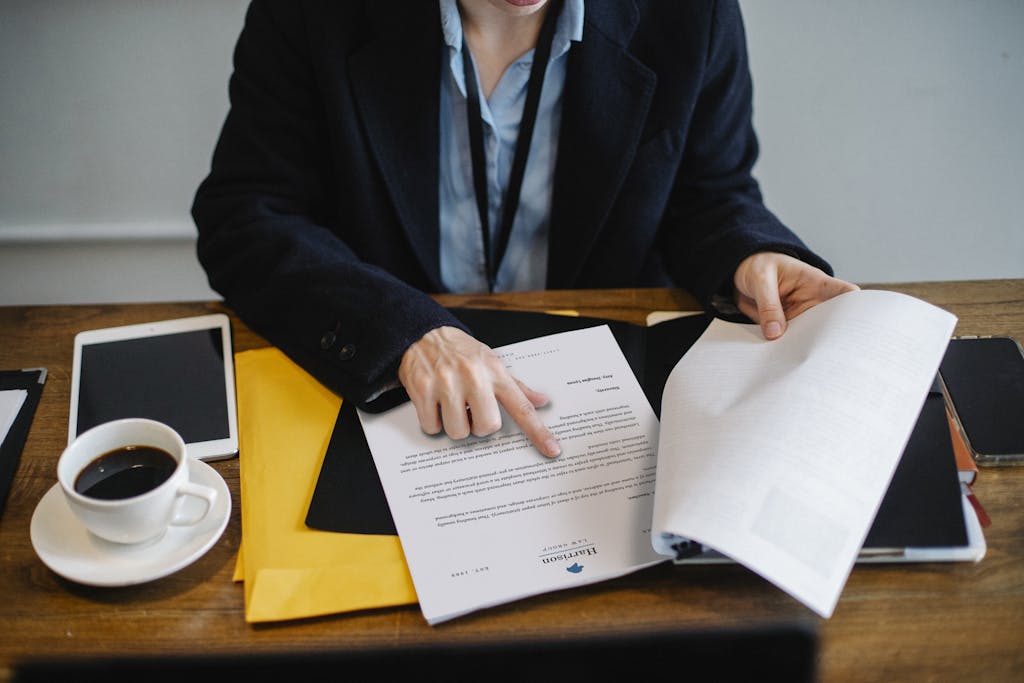 High angle of crop faceless woman in formal wear sitting at wooden table with tablet and coffee and reviewing a redline contract.